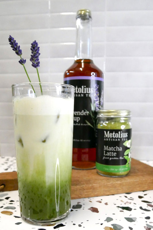 Iced lavender matcha latte drink showing oat milk layered with matcha latte mix in a tall glass with drizzle of lavender syrup and a sprig of lavender